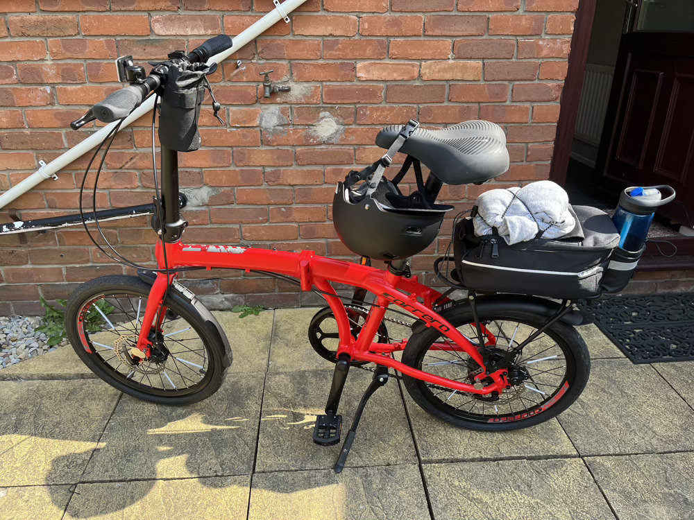 My little red folding bike, leaning on its kickstand. The black spokes on the wheels have white spoke reflectors attached. My helmet is dangling from its strap on the saddle. There’s a small bag attached to the ‘inside’ of the handlebars and a rear rack bag that’s loaded up