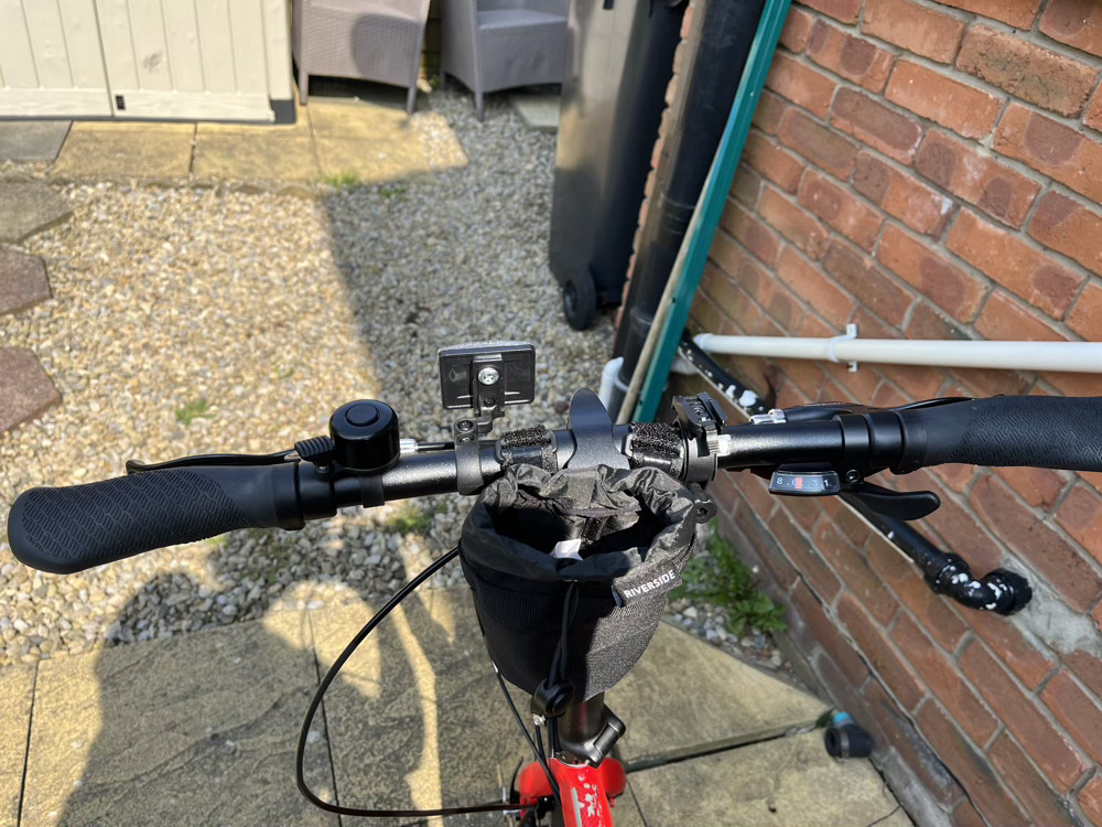 A view of the handlebars. The bars themselves are black, as are the accessories that are attached. From left to right: there’s a bell nearest the left grip, then a front reflector screwed on near the middle. A small water bottle bag is velcro’d to the middle of the bars and the post. To the right of this is a bracket for attaching the front light, then the gear shifter is nearest to the right grip