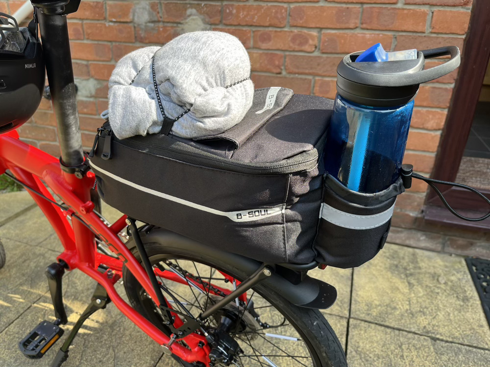 A close up of the rear rack and rack bag. The bag is mostly black with a small silver reflective strip. It looks quite full. On top, a light grey hoodie is fastened by elastic straps. There’s a small water bottle pocket on the back, containing an empty blue water bottle