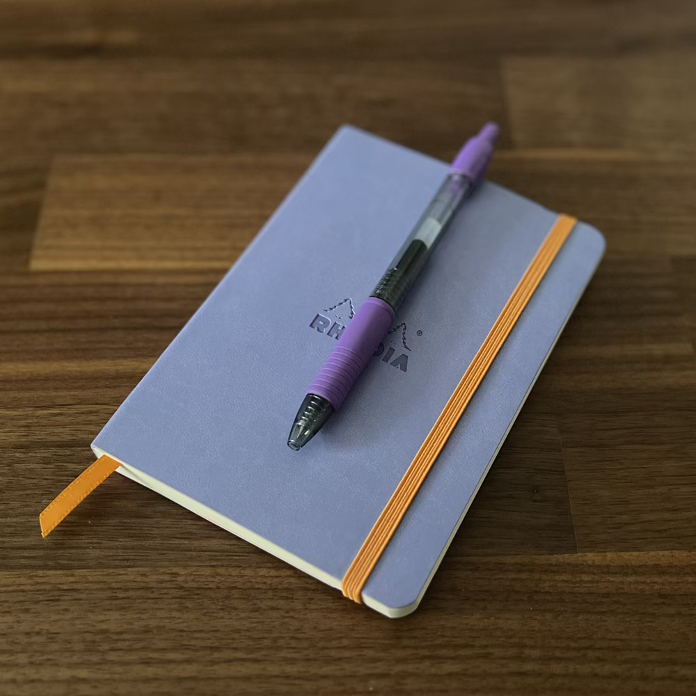 A light purple A6 Rhodia notebook lying on a wooden surface. A purple pen is clipped to the outside of the front cover