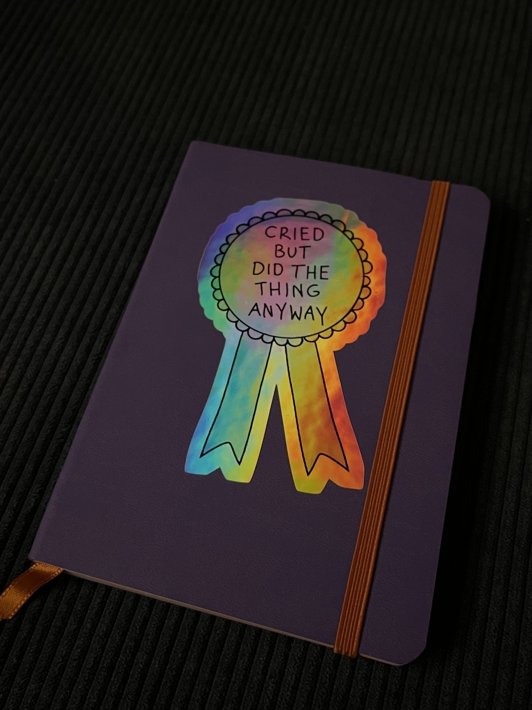 A light purple A6 notebook. A holographic sticker in the shape of a rosette, reading ‘cried but did the thing anyway’ is stuck on the front 